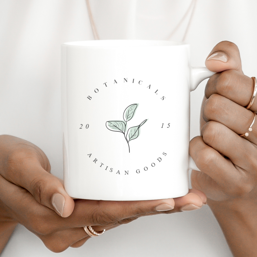 "Botanicals Artisan Goods" curved text design on white coffee cup with artsy leaf graphic.