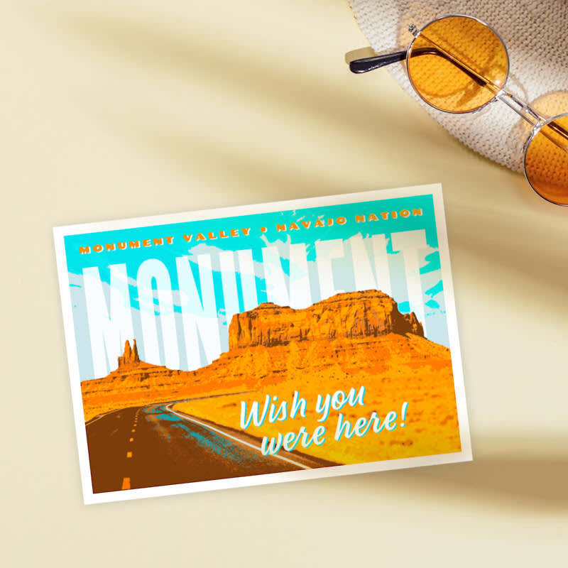 Bright postcard with blue sky, white clouds, and orange desert rocks alongside a highway road. Text reads in big white font: MONUMENT, with light blue shadowed font stating, "Wish you were here." Beige behind postcard with orange circular sunglasses.