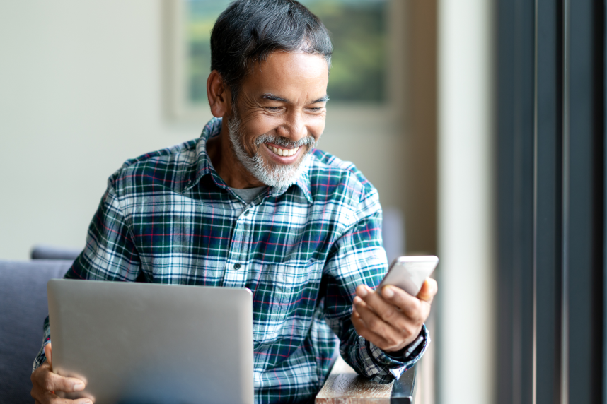 Male holding a laptop and smartphone, smiling at screen. 