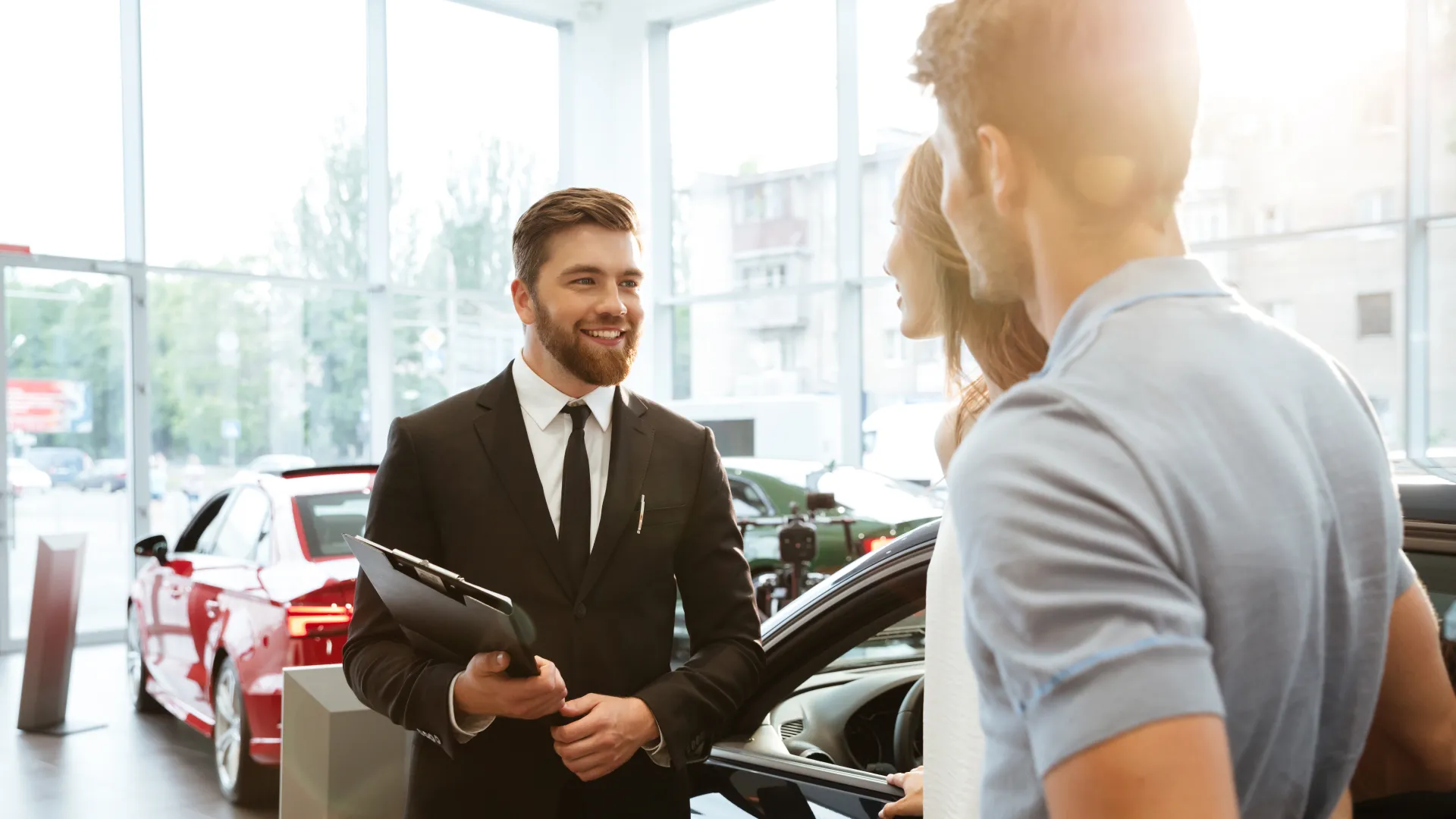 A photo of a man in a suit holding a clipboard smiling at a couple who are looking at cars in a dealership showroom