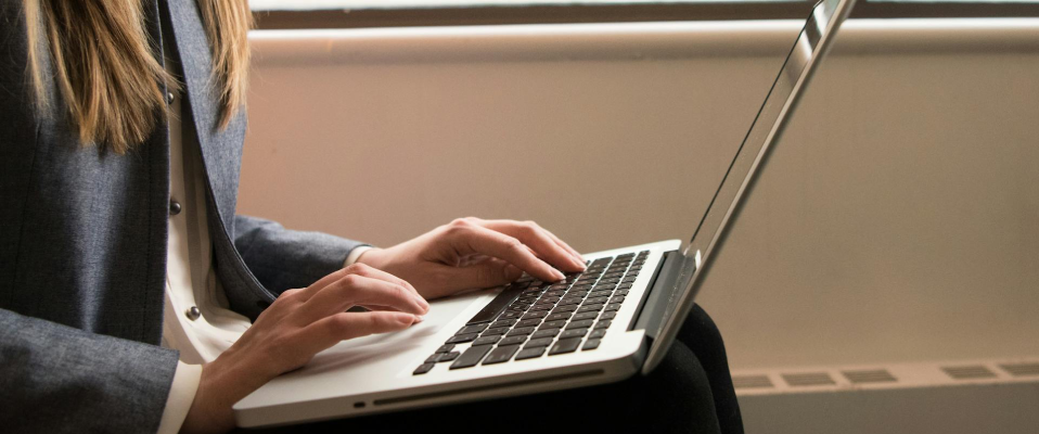 A side-view photo of a woman in a suit jacket typing on her laptop, which is in her lap. Her face is not visible, and the frame is centered on the keyboard.