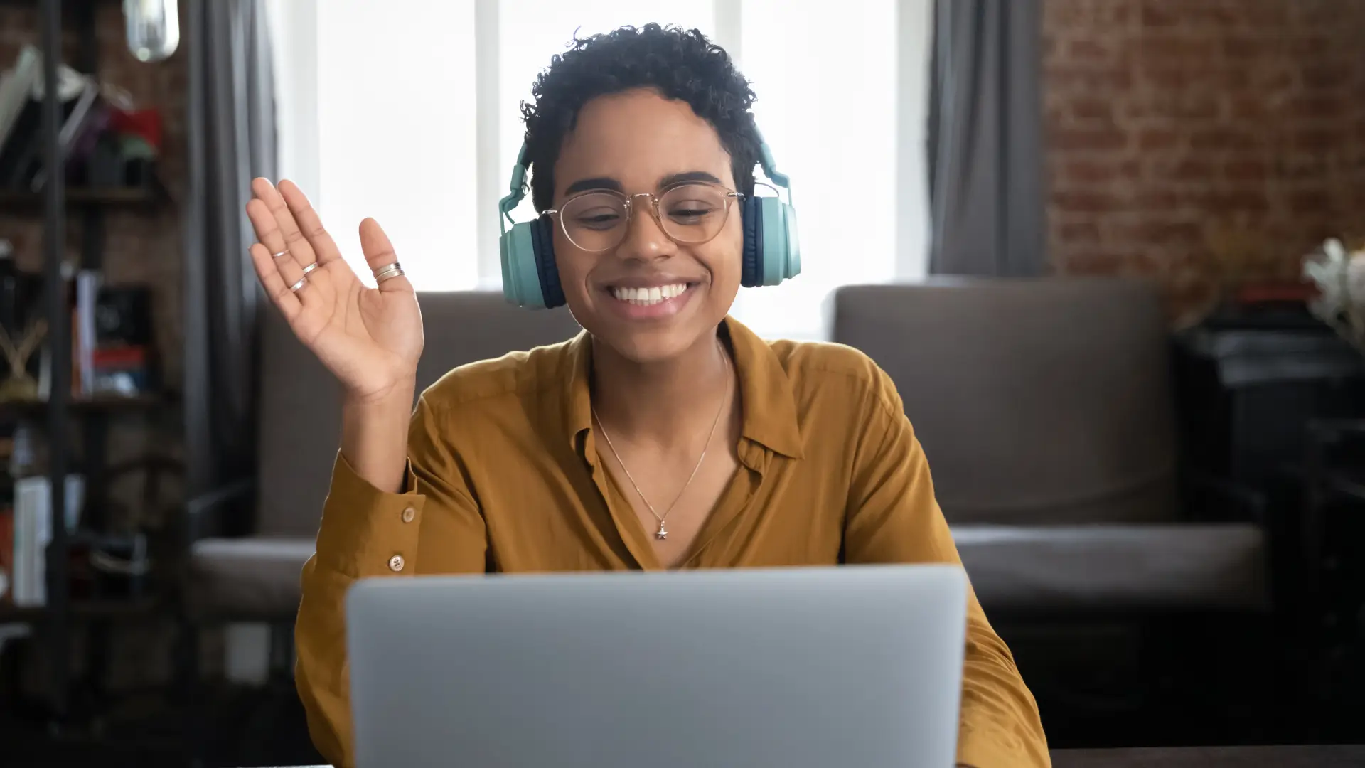 A photo of a young woman wearing headphones smiling and waving into her laptop camera. She is sitting in front of a brick wall with a bright window in what looks like a home office.