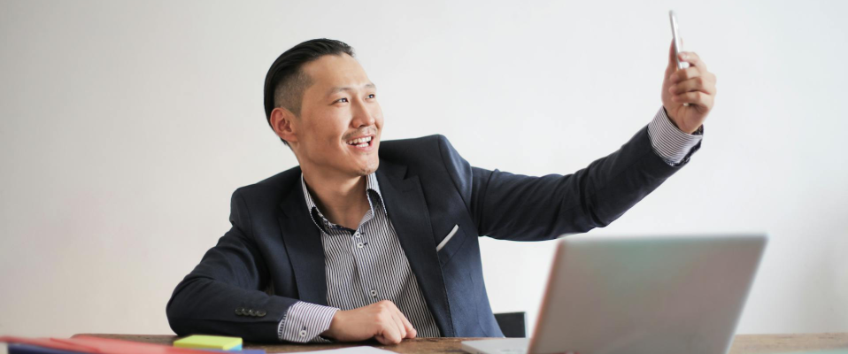 A man in a suit sits at his desk in front of an open laptop and some papers. He is holding his phone up and smiling into the phone camera.