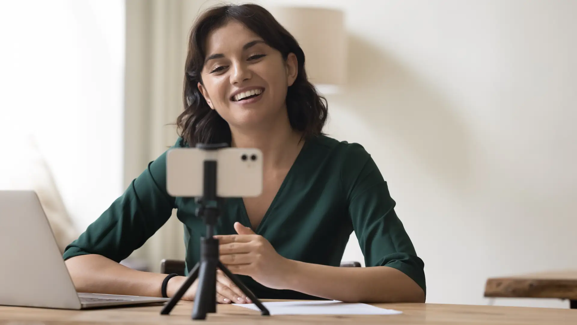 A woman sitting at a desk next to her laptop smiles into her phone, which is held up by a tripod so she can record a video.