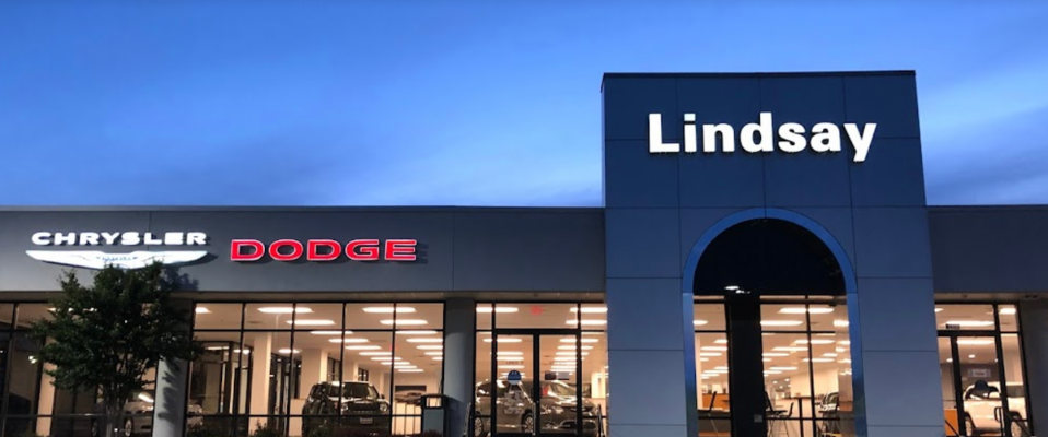 A photo of a car dealership at night, the signs for the Chrysler and Dodge logos are lit, and there is a large arch with the name of the store, Lindsay, above the entrance