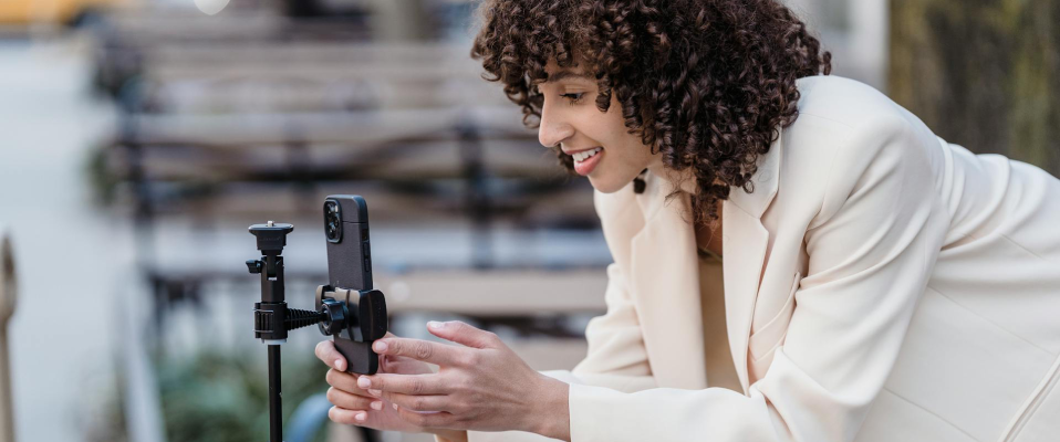 A woman in a white suit smiles into her phone which she is setting up on a camera stand to start recording a video.