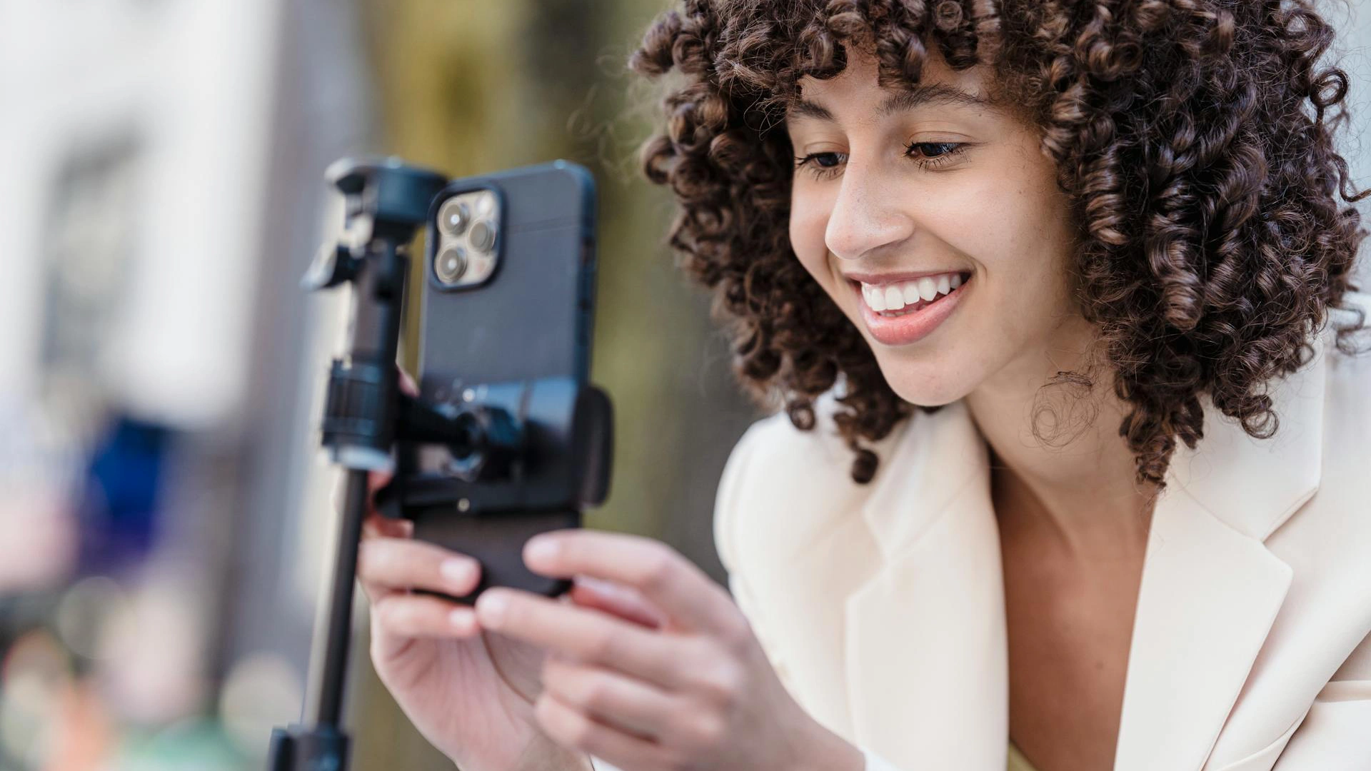 A woman in a white suit smiles into her phone which she is setting up on a camera stand to start recording a video.