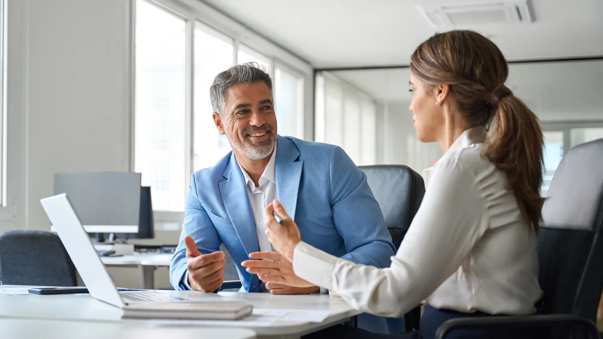 A man and a women sit at a table in a white board room wearing business casual clothes. They smile in front of a computer while they speak.