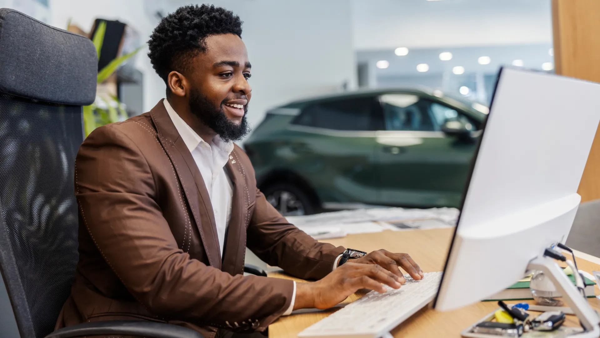 A man in a suit sits at a dealership and types on the compuyer with a car in the background