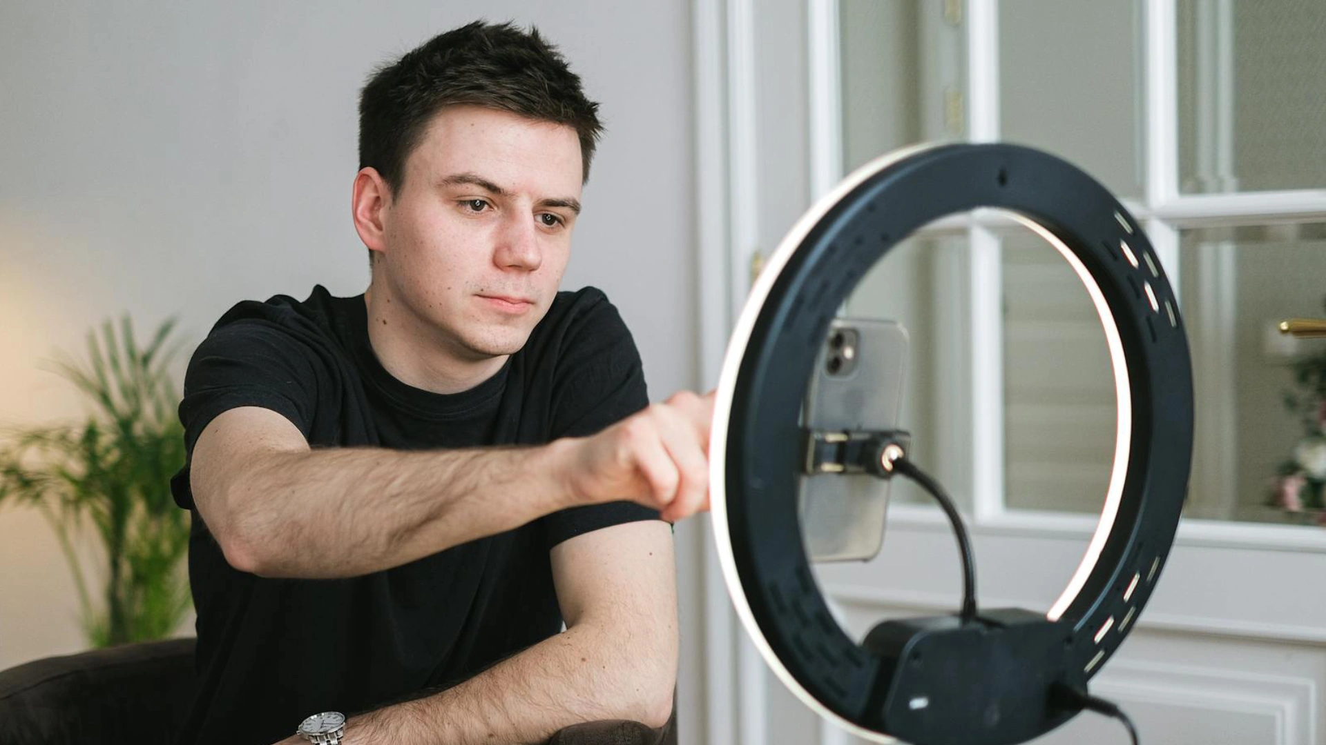 A man in a black tshirt sits in a white living room in front of a phone mounted to a ring light. He is reaching out and clicking the screen.