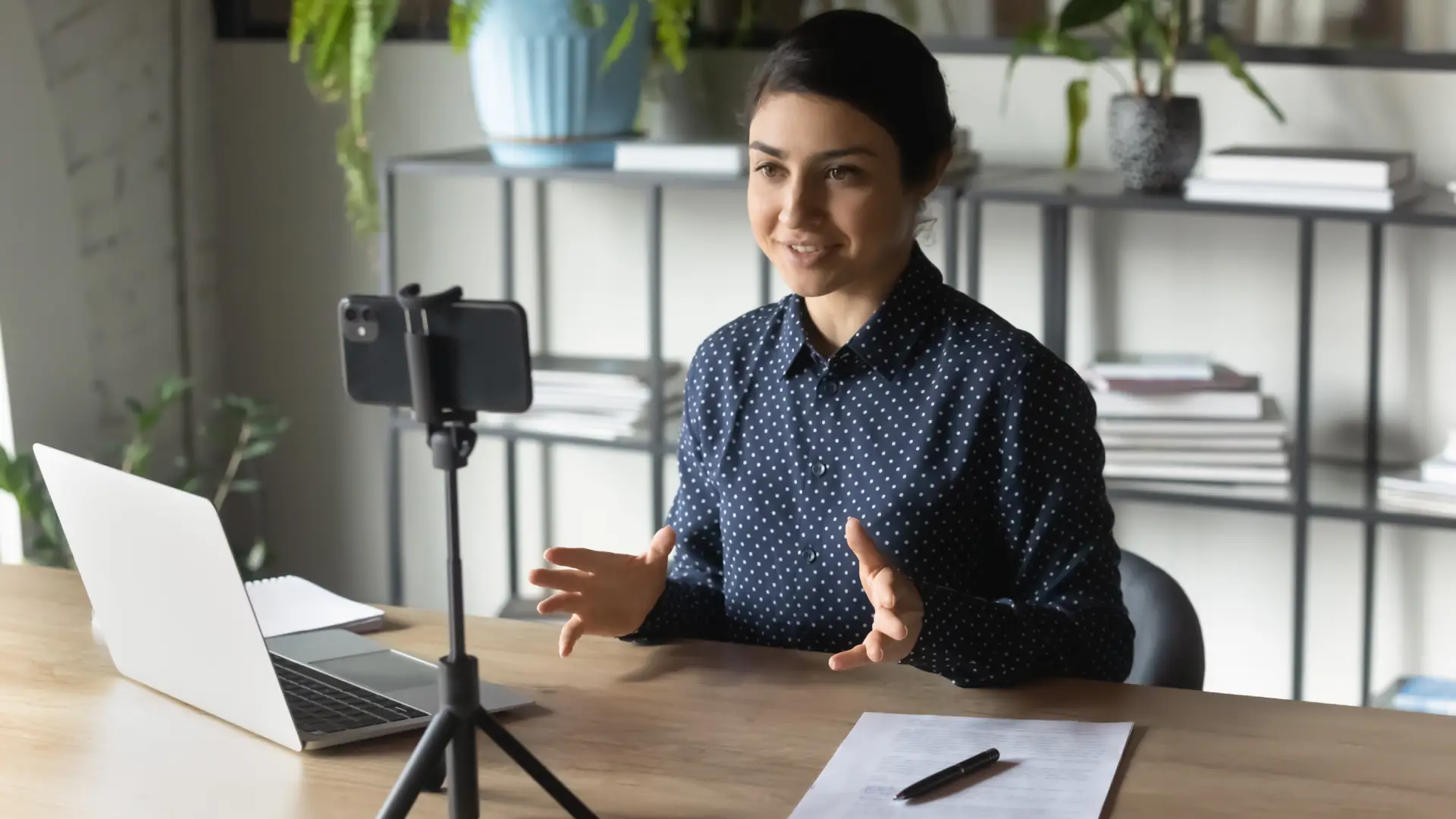 A photo of a woman at her desk in front of a laptop. She is smiling and speaking into her cell phone camera, which is held up by a small tripod.