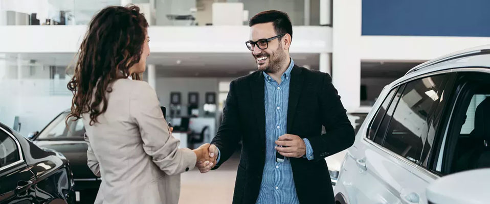 A man and woman shake hands in front of a white car inside a dealership