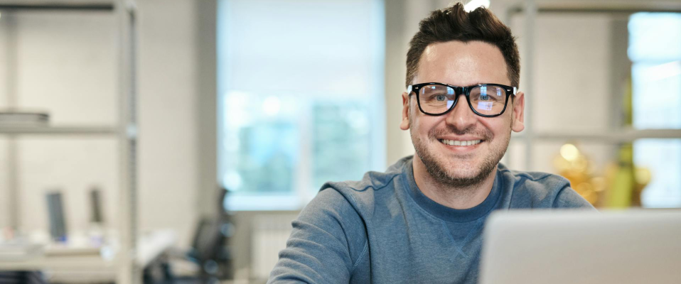 A photo of a man wearing glasses sitting in an office. There is a laptop screen in front of him as he smiles into the camera.