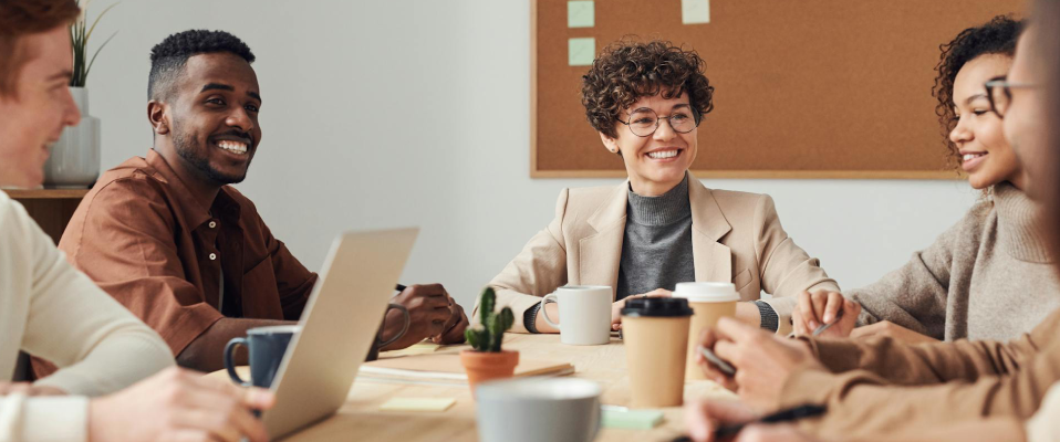 A photo of people in business casual attire sitting around a table smiling and discussing. There are coffee mugs, notes, and a laptop on the table as they work.