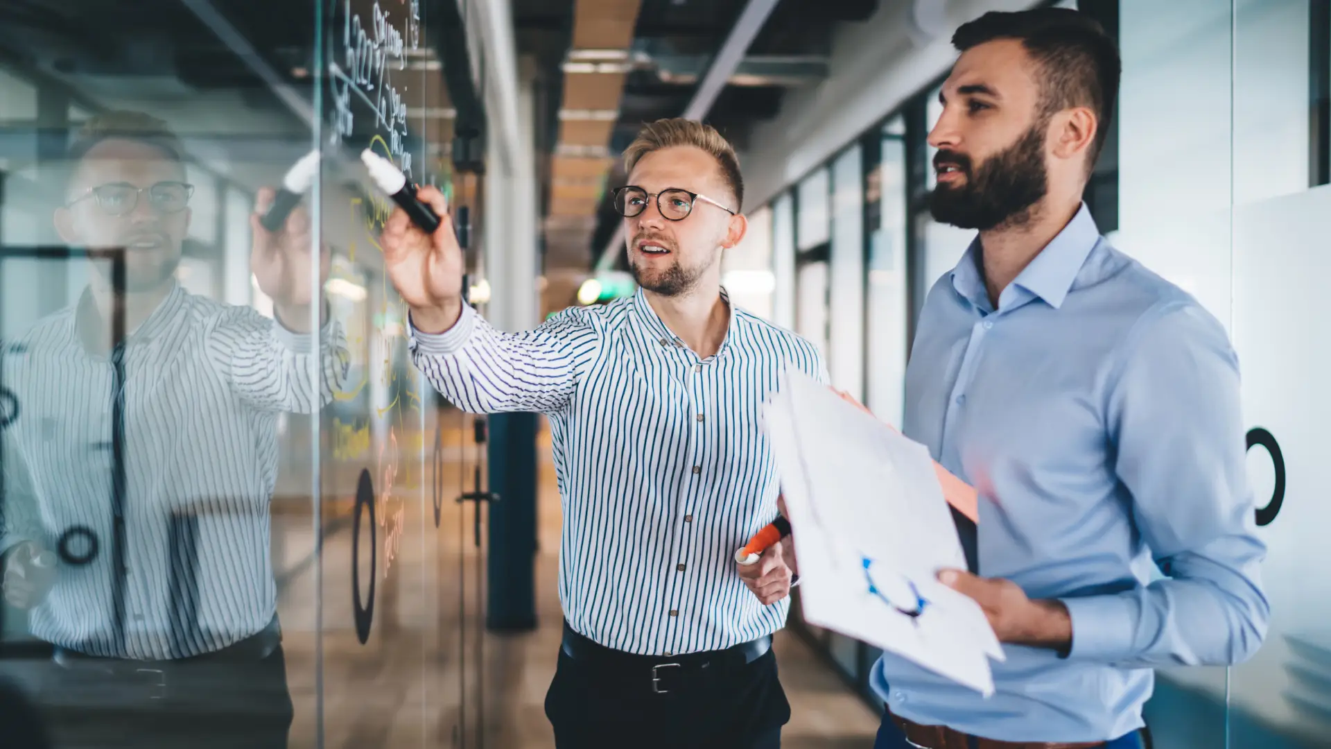 A photo of two men dressed in business casual standing side by side in front of a clear wall. One is holding papers with charts, and the other is writing notes on the wall.