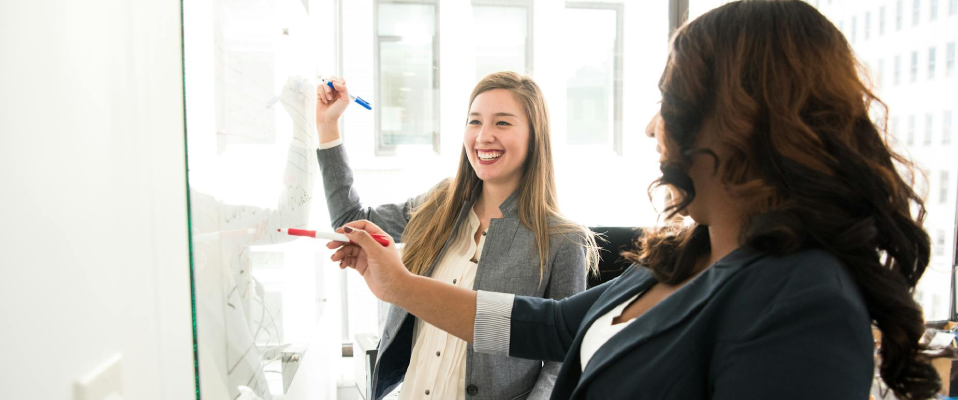 Two women stand at a whiteboard, writing ideas in a brightly lit office. One is turned to the side, and the other is smiling in the direction of the camera.