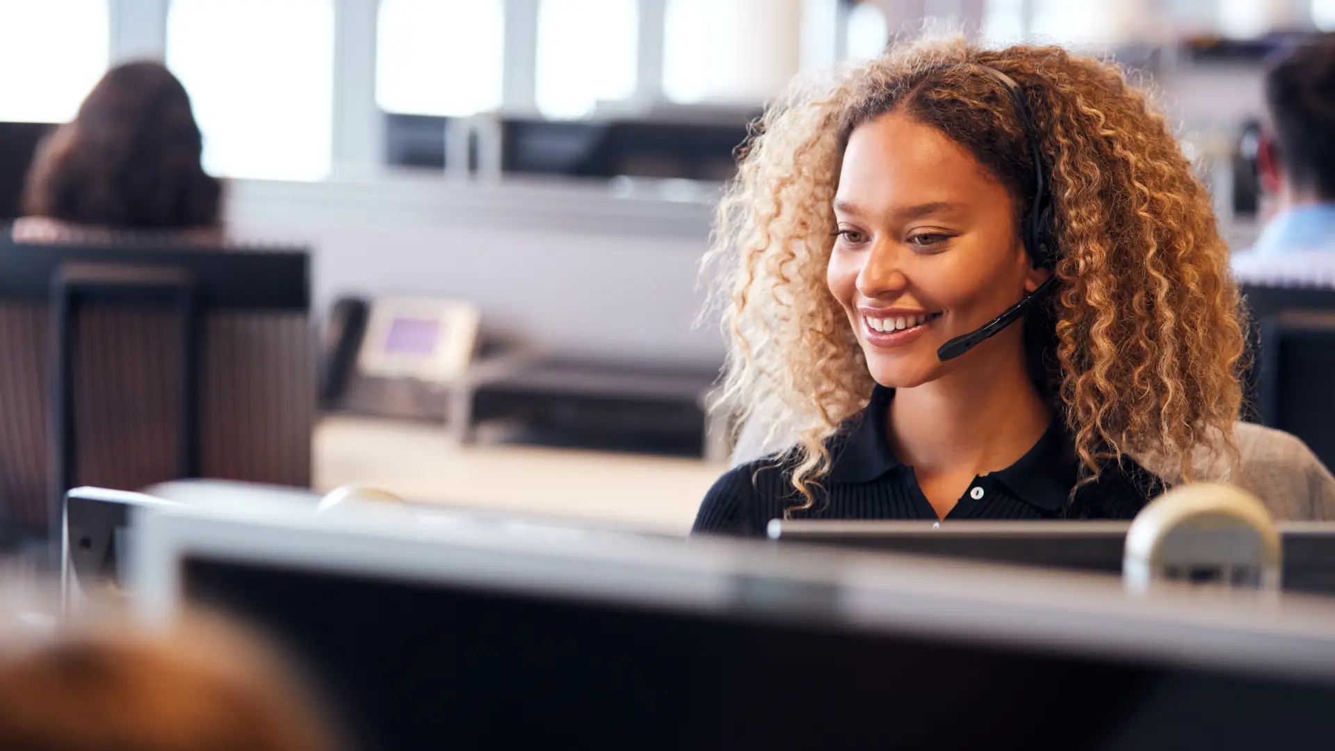 A woman with curly hair and a button-up shirt sits at a computer wearing a headset and smiling. She appears to be in an office or call center, speaking with potential customers.