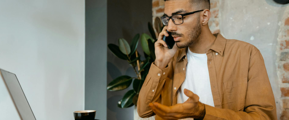 A man sitting at a text talks on his phone as he looks at a computer screen