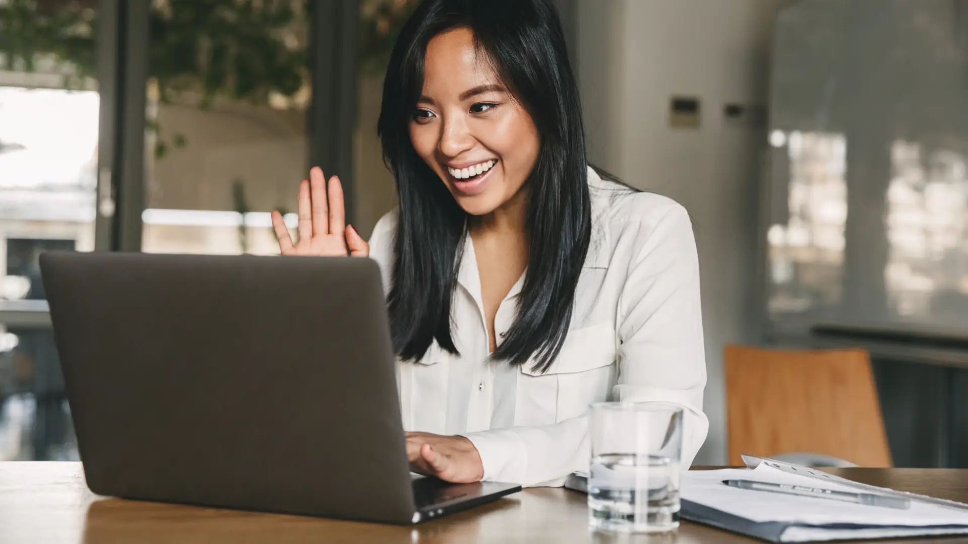 A woman in a button-up shirt sits in an office and smiles and waves into her laptop camera as she records a video