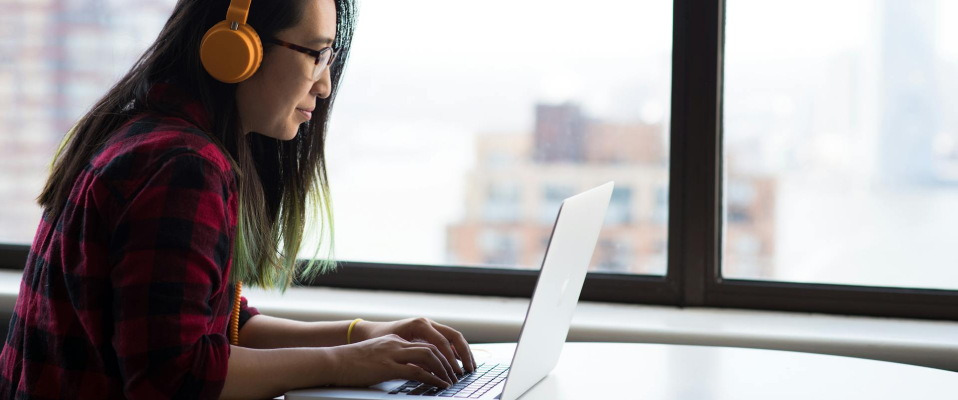 A photo of a young woman wearing headphones typing on a laptop at a table that overlooks a city outside