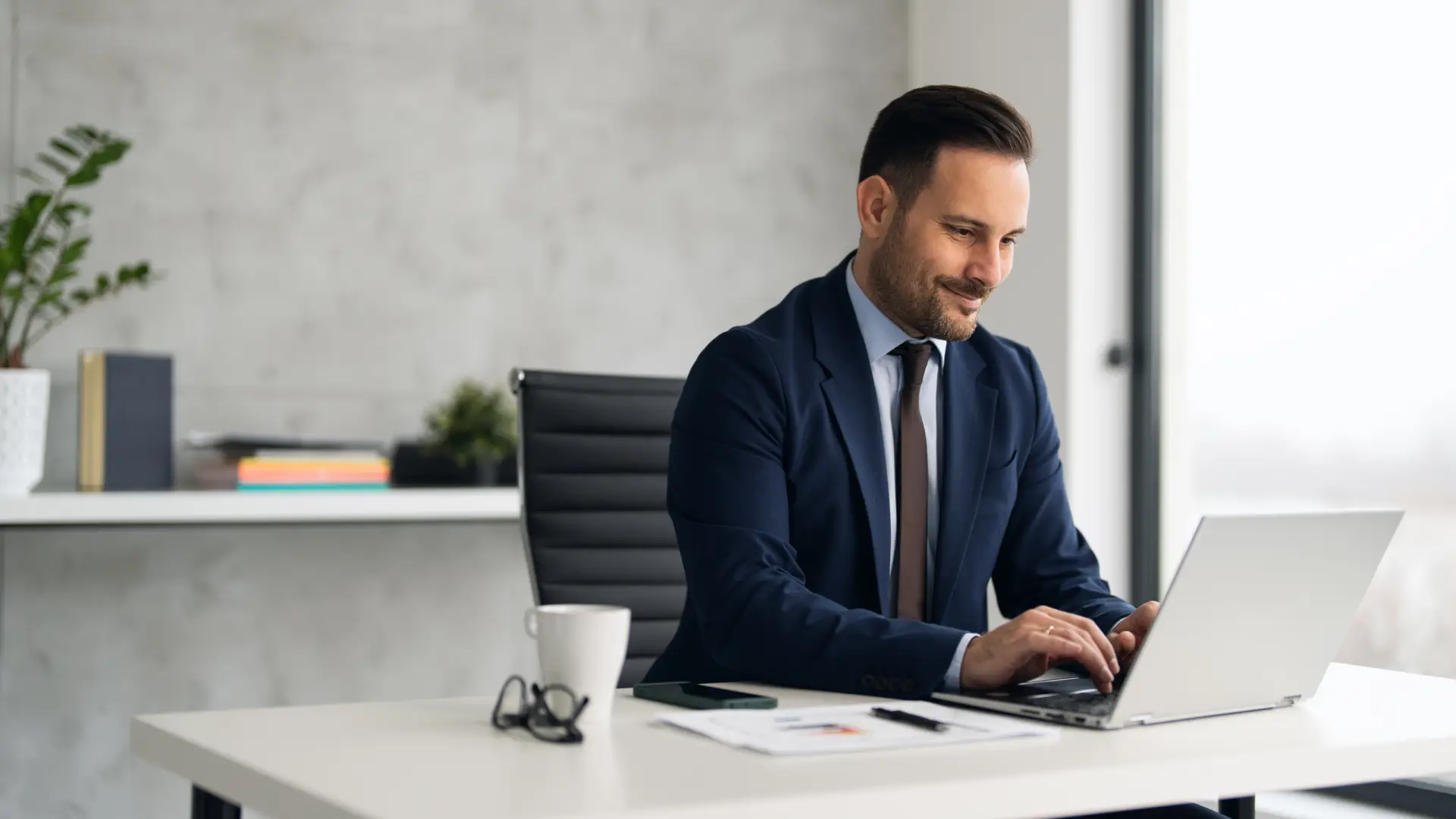 A man in a suit sits at his desk in the office and types on a laptop