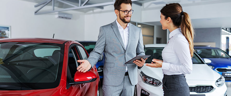 Smiling car seller standing in car salon with customer and showing around cars on sale.