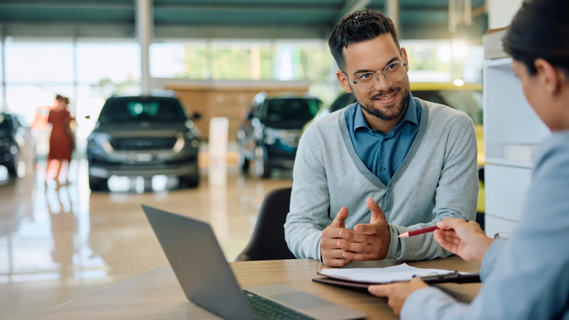 A man in a collared shirt and cardigan sits across from another man outside the frame discussing details written on a clipboard. Behind them, you can see a dealership showroom.