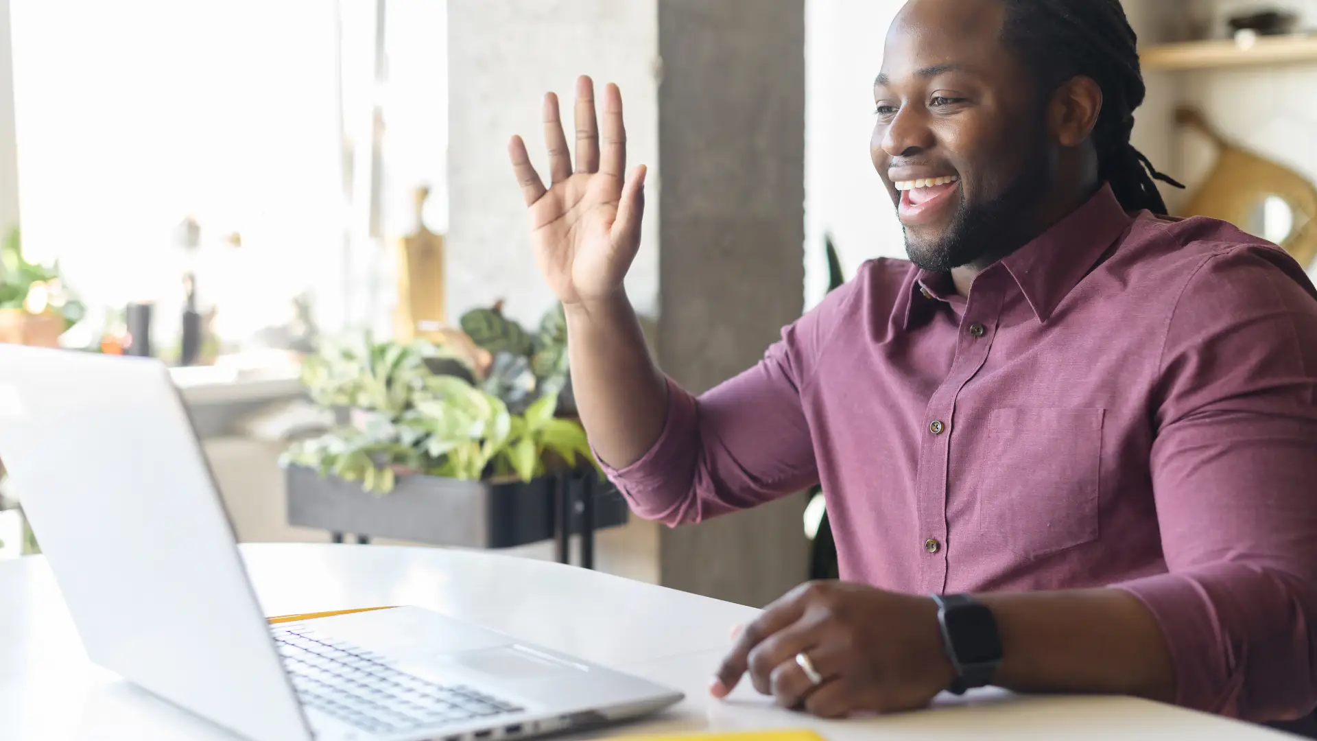 A photo of a man in a button-up shirt sitting at a desk in an office. He is smiling and waving into the webcam on the laptop in front of him.