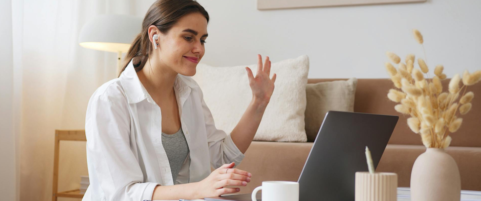 A woman sitting at a table wearing earphones smiles and waves at her laptop computer screen