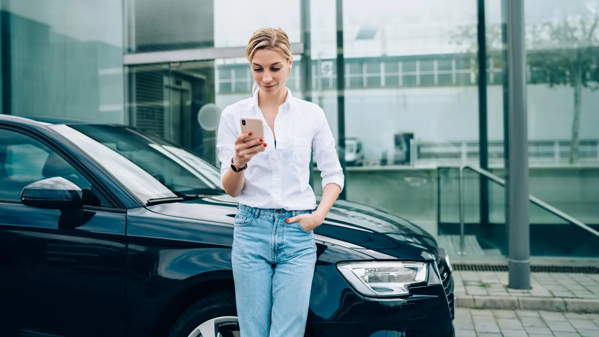 A woman in a button up and jeans stands with her back to a car while she looks at her phone, smiling