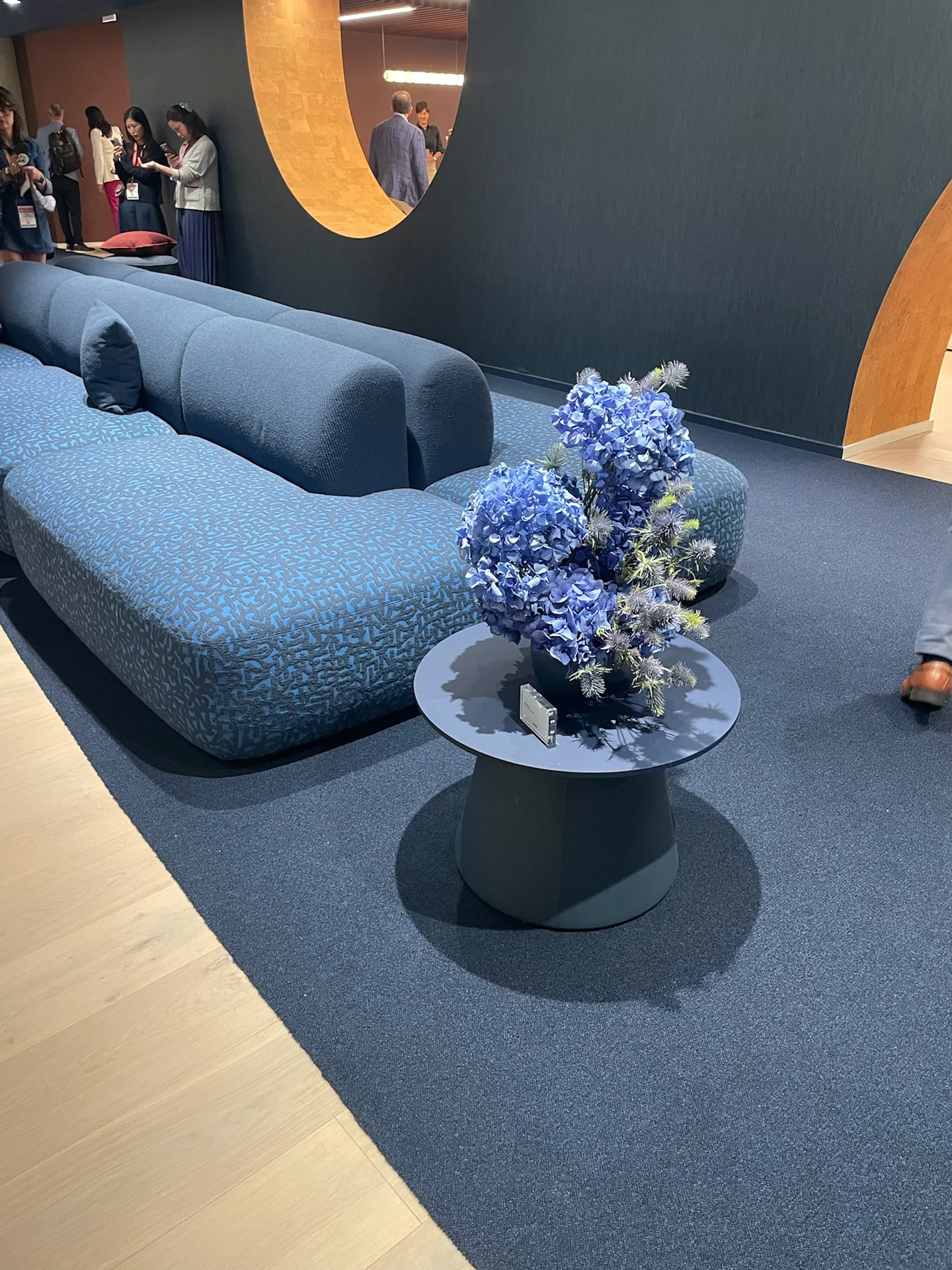 Curved blue upholstered seating with textured fabric in a modern lounge setting, paired with a round table and floral arrangement on a coordinating blue carpet