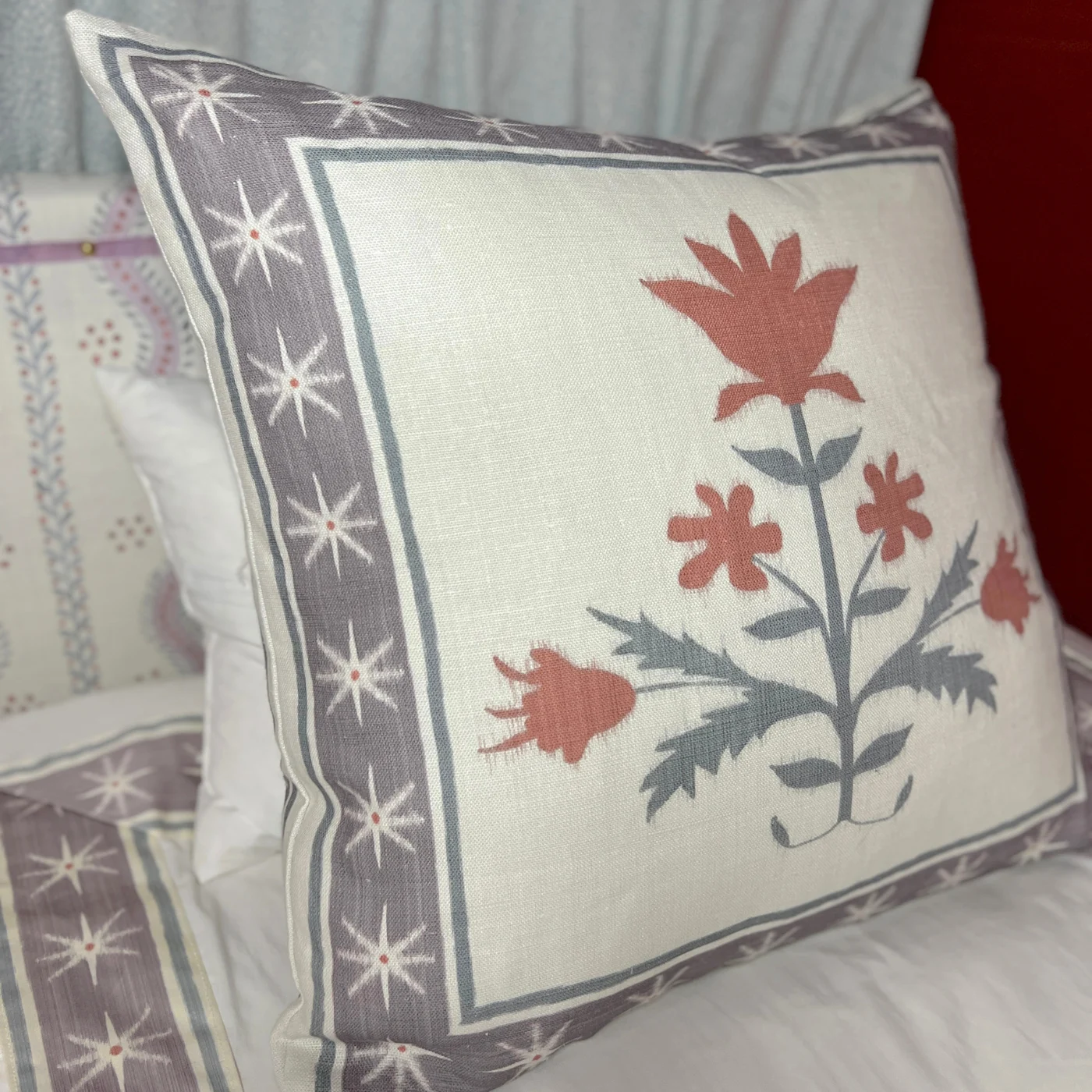 A white pillow with bands just inside each of the four border edges featuring a thick purple stripe with white stars. The stripe has thin blue stripes on either side of it. Red flowers with green stems are at the center of the design. The pillow is shown