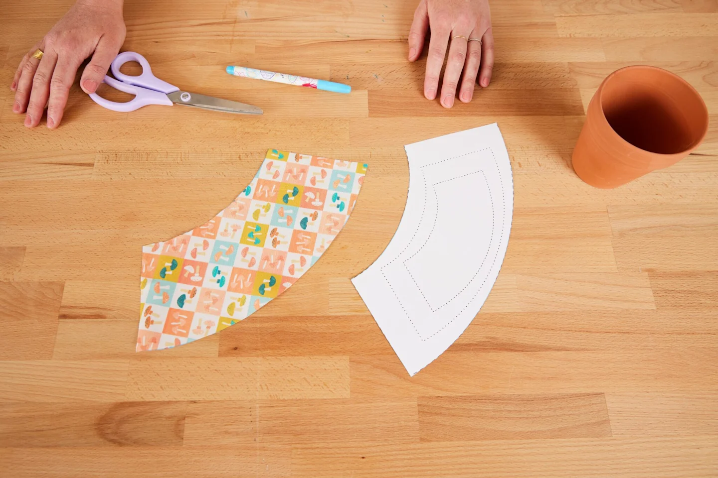 A cut-out pattern piece in both paper and fabric lay on a wooden table. They are fit to cover a 4” (10.2 cm flower pot).