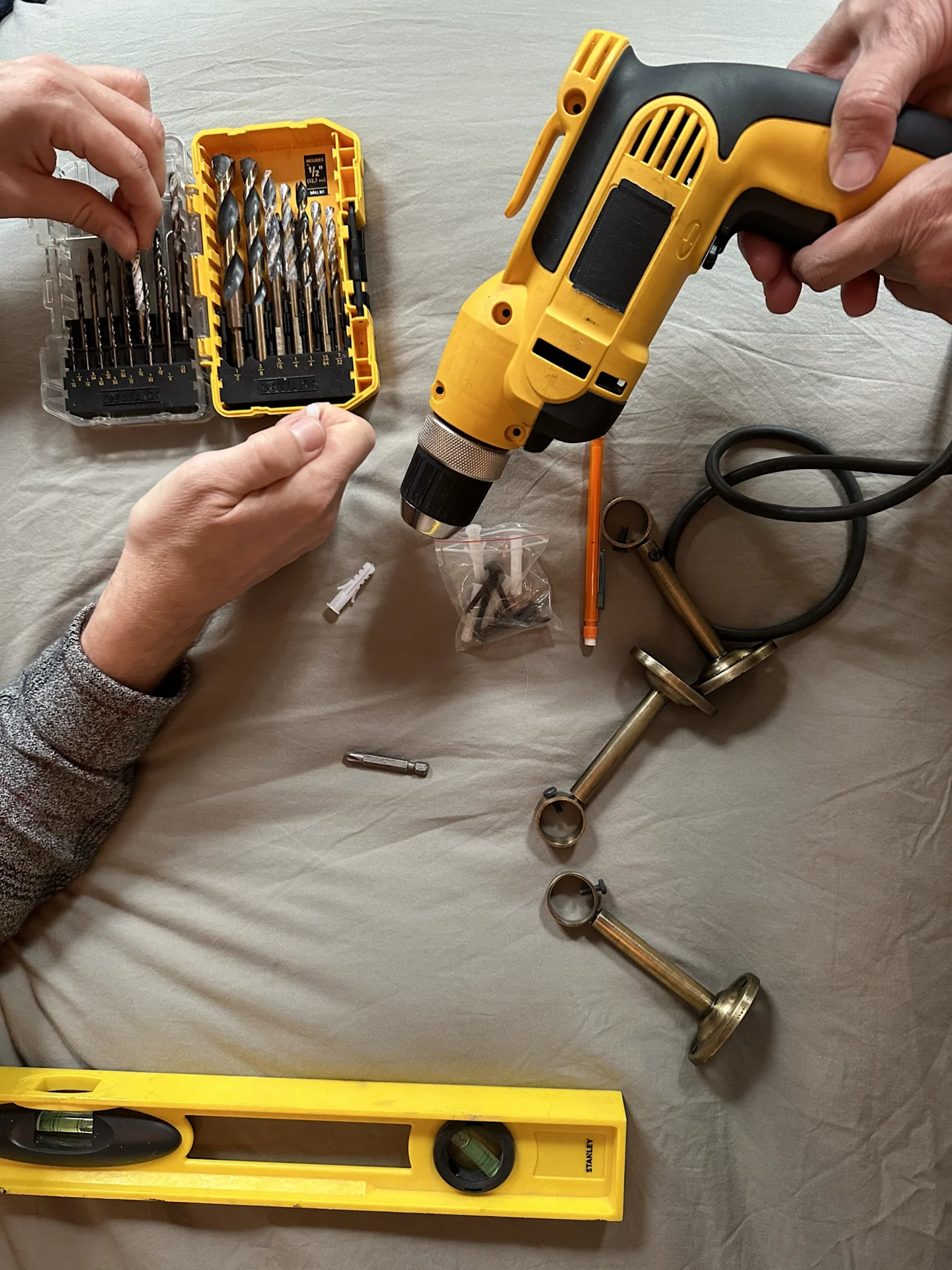 A yellow spirit level partially extends into the bottom left of the photo. Curtain installation hardware, drill bits, a pencil and a drill lay on a light gray sheet on a bed. In the top left, a hand is going through a container of drill bits. In the top r
