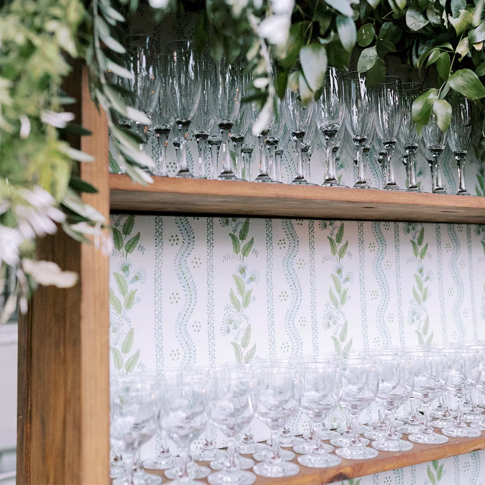 Bar at a wedding reception with a shelf with glassware and floral striped wallpaper behind it.