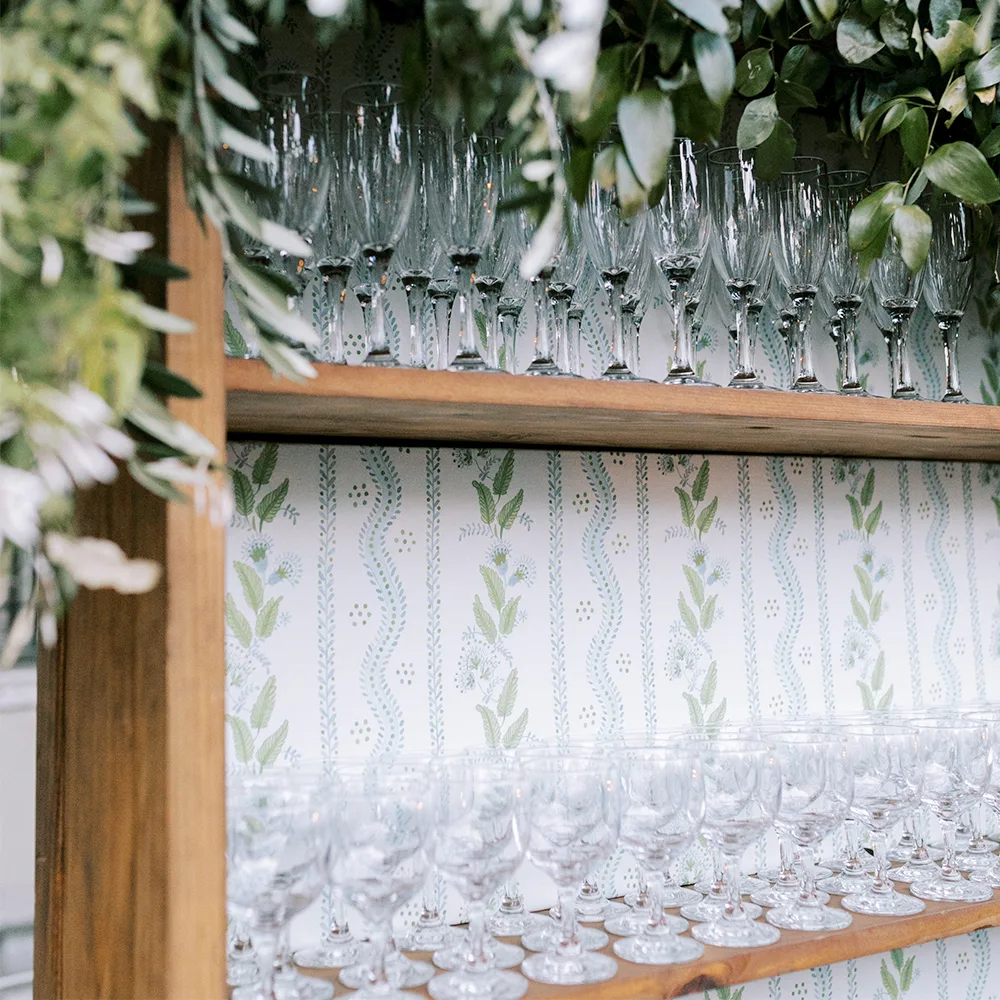 Bar at a wedding reception with a shelf with glassware and floral striped wallpaper behind it.