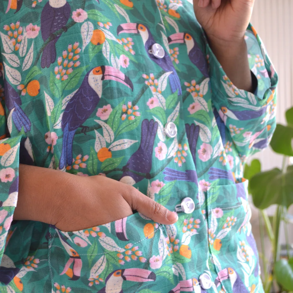 A close up of Rumana’s pajamas showing the top’s front buttons and pockets. Rumana’s left hand is in her pocket. The pajamas have a tropical print featuring toucans, pink and orange flowers and a bright blue background. Behind Rumana is a gray couch and a