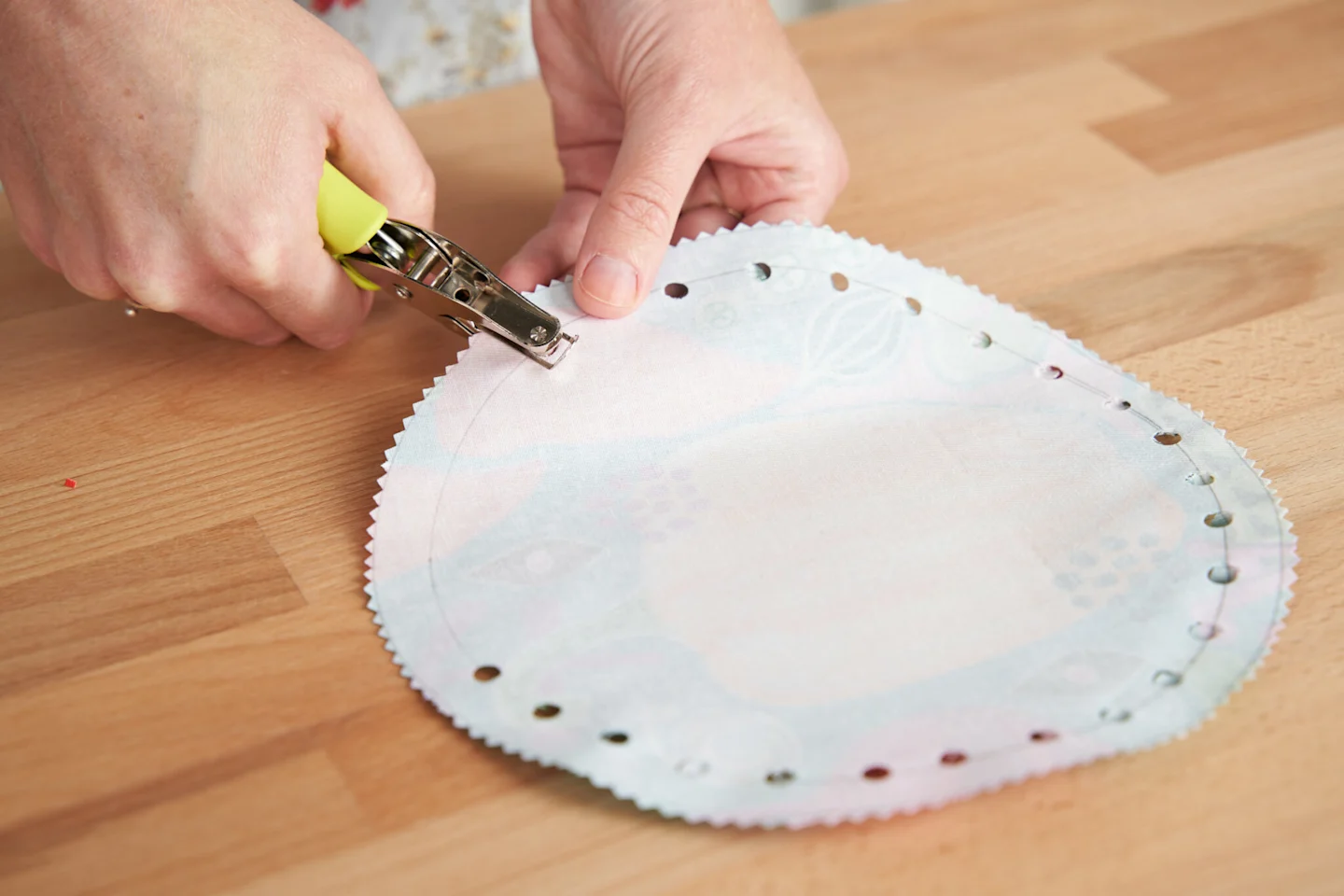 A person is using a hole punch to punch holes around the first, smaller circle drawn onto the wrong side of fabric with a compass.