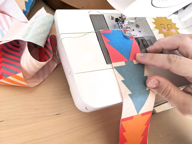 A 2”-wide strip of fabric with a Christmas tree repeating design in jewel tones has been placed under the needle of a sewing machine loaded with blue and yellow thread. The needle has been placed 1/4" in from the right-hand side of the fabric strip.