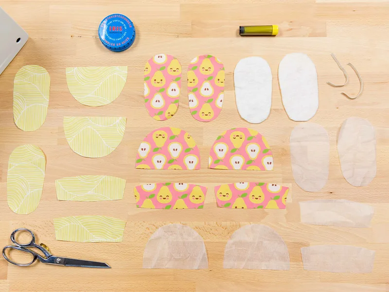 Cut out pieces of fabric and interfacing to make baby shoes lay on a wooden surface with a pair of fabric scissors and a blue tin of pins.