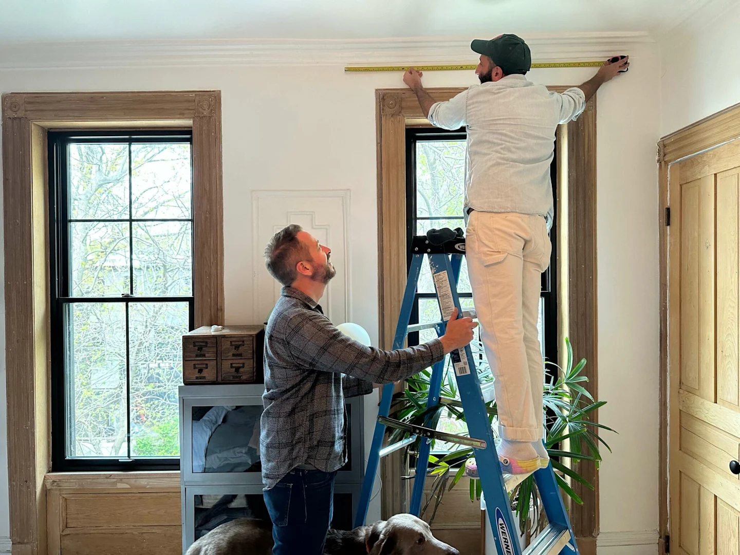 A man is standing on a ladder to measure the top of a window. He holds a yellow measuring tape along the top of the window. Another man is holding the ladder to make sure it doesn’t tip over.