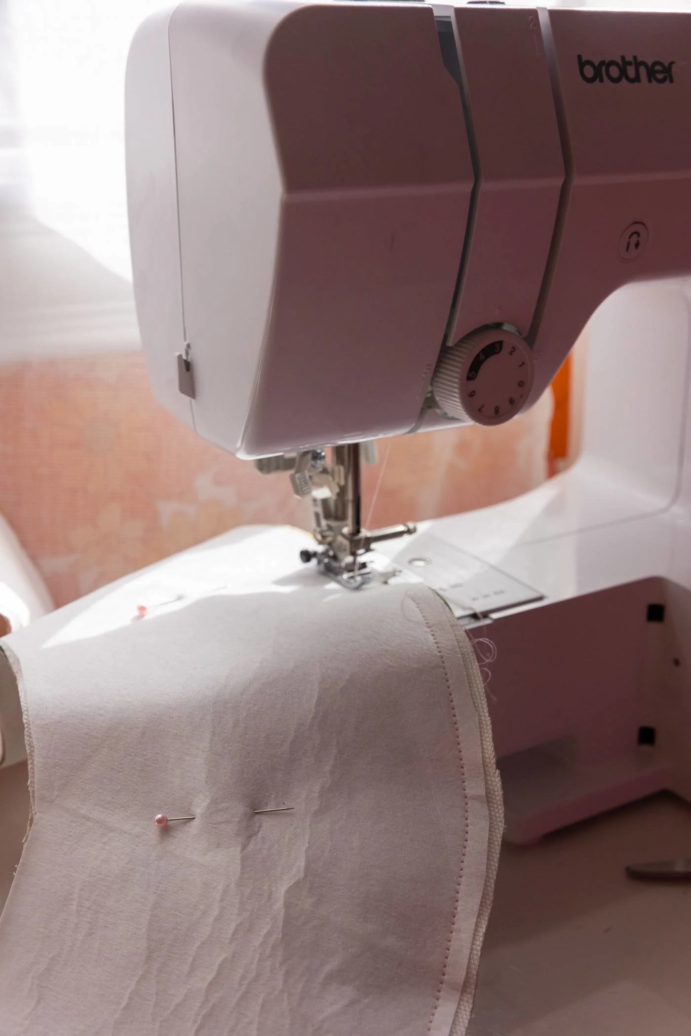 A sewing machine stitches the edge of the brim. Sewing pins are distantly placed in the middle. Out of focused is a sheer curtain filtering the sunlight from a window on a wall covered in a retro flower wallpaper.