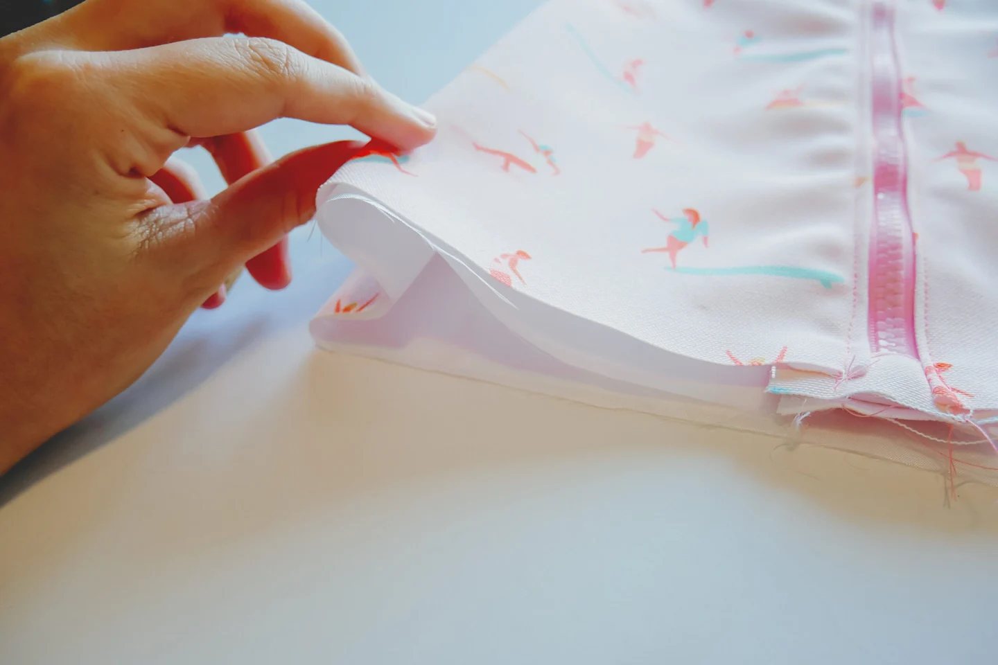 A fabric rectangle featuring a design with small female surfers surfing through a pink background lays on a white surface. A pink zipper has been stitched up the middle. A hand is folding the sides of the fabric back in on themselves, making a sideways M