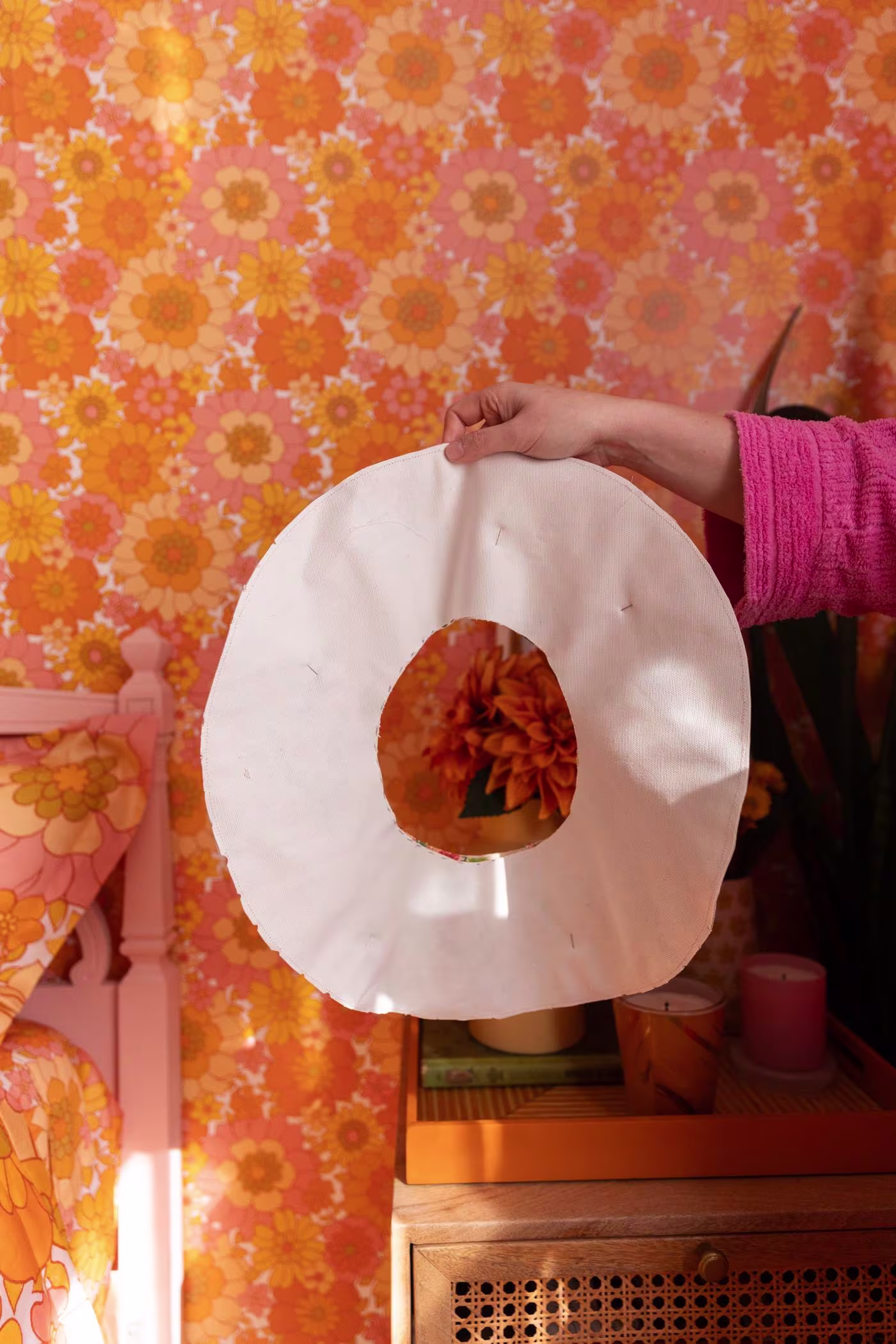 Keiko holds up the brim. Small notches are around the outer edge of the brim. Retro floral wallpaper and bedding to match are seen from the left corner. A desk holding candles and flowerpots is behind Keiko to the right.