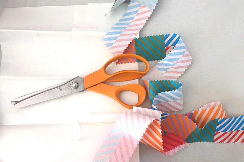 A long strip of 2”-wide cotton fabric featuring a design with small squares of coordinating colors in varying jewel tone pairings has been cut out with pinking shears. It is laying on a white surface. A pink or orange-handled pinking shears is laying on t