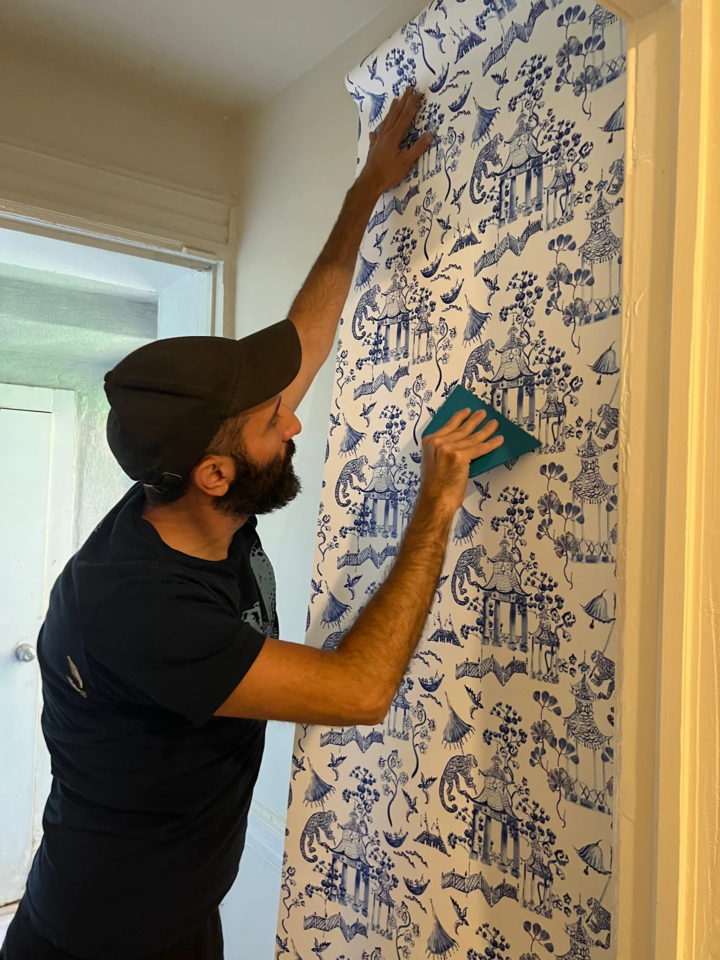 A man is installing wallpaper with a blue-and-white chinoiserie design on the walls of hallway.