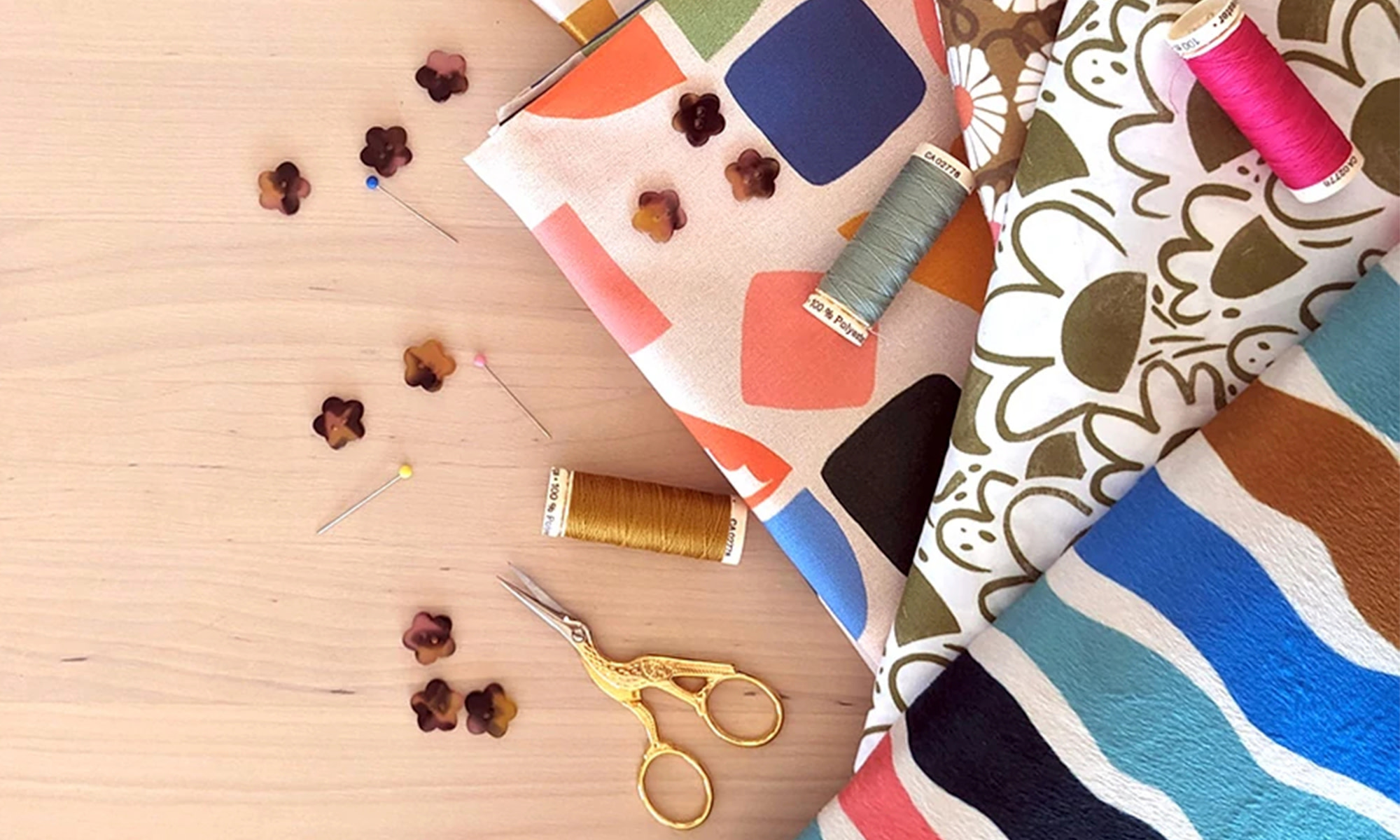 Fabrics, thread, embroidery scissors buttons and pins on a wood table.