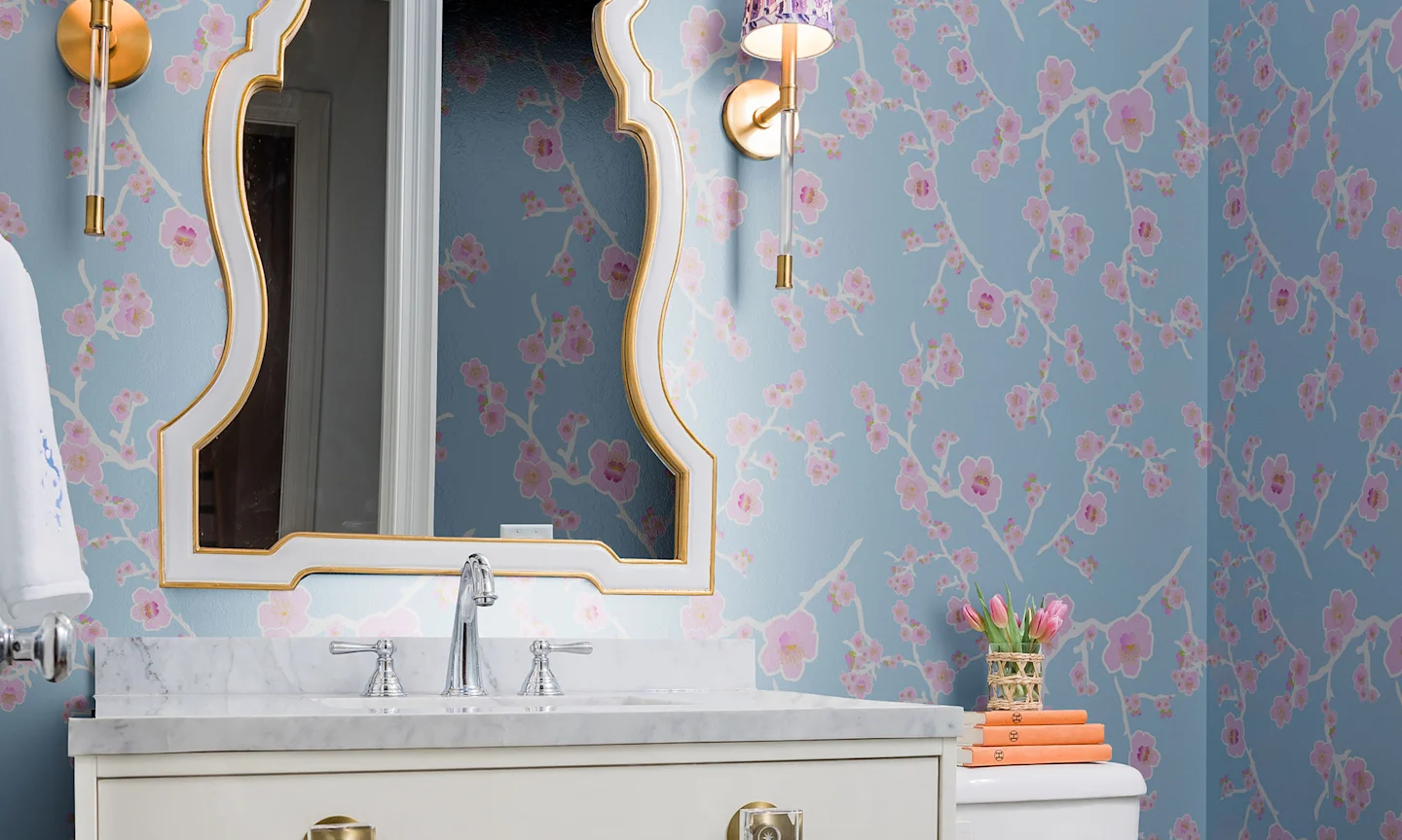 A bathroom with blue and pink floral wallpaper, a marble countertop with a sink, a gold-framed mirror, and two wall-mounted lights.