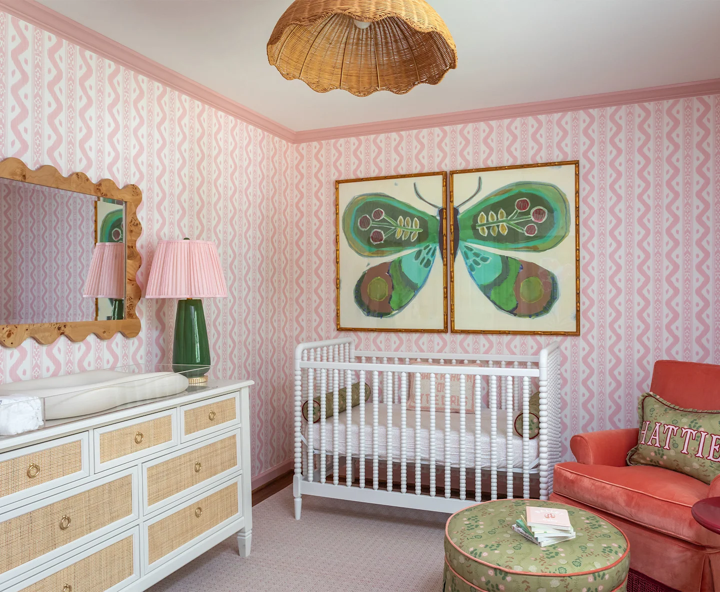 Nursery featuring pink patterned wallpaper, a white crib, cane-front dresser, scalloped mirror, butterfly artwork, and a coral armchair.