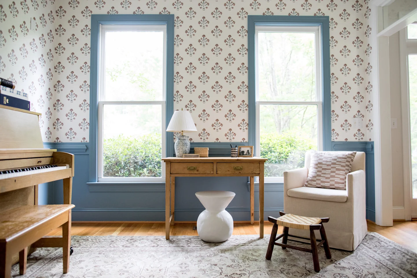 Katherine’s same sitting room after it has been madeover. A thin wooden table is underneath two large windows with two front drawers. Blue paint has been added around the windows and under the chair rail. Wallpaper has been added over the chair rail with
